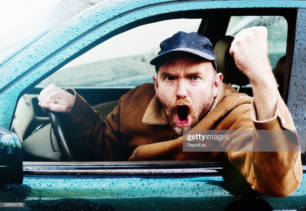 A man shakes his fist furiously outside of his car.