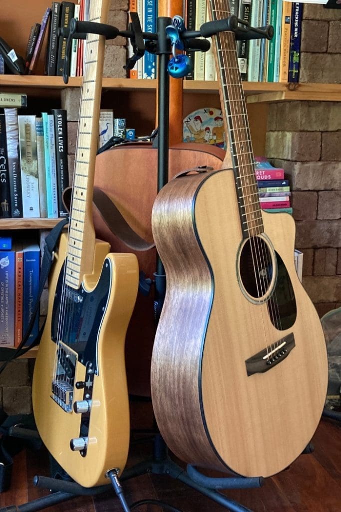 Three guitars resting on a guitar stand in front of a shelf of books